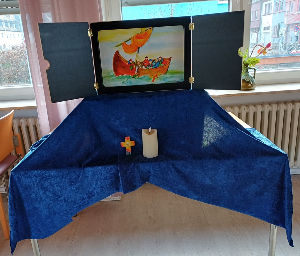 Auf einem Tisch ist ein mit einem blauen Tuch überdeckter Sockel auf dem ein Kamishibaitheater aufgebaut ist. Davor steht ein Kreuz und eine Kerze