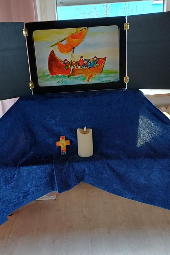Auf einem Tisch ist ein mit einem blauen Tuch überdeckter Sockel auf dem ein Kamishibaitheater aufgebaut ist. Davor steht ein Kreuz und eine Kerze