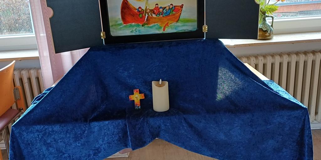 Auf einem Tisch ist ein mit einem blauen Tuch überdeckter Sockel auf dem ein Kamishibaitheater aufgebaut ist. Davor steht ein Kreuz und eine Kerze