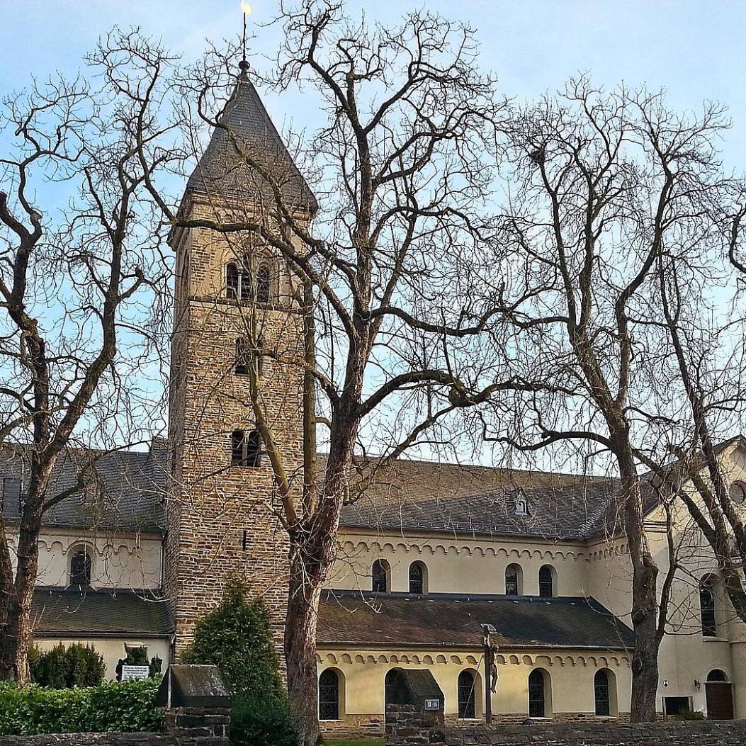 Kirche St. Laurentius von außen, von der Straßenseite her, die großen Kastanien davor haben keine Blätter. Blauer Himmel.