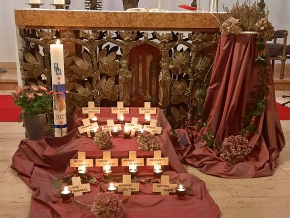 Vor einem Altar und der Osterkerze ist ein rotes Tuch ausgebreitet, darauf sind mehrere Holzkreuze ausgelegt mit Namen der Verstorbenen des letzten Jahres und Kerzen