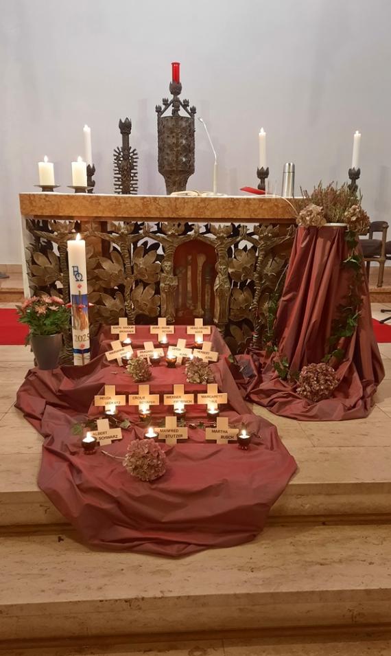 Vor einem Altar und der Osterkerze ist ein rotes Tuch ausgebreitet, darauf sind mehrere Holzkreuze ausgelegt mit Namen der Verstorbenen des letzten Jahres und Kerzen