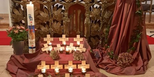 Vor einem Altar und der Osterkerze ist ein rotes Tuch ausgebreitet, darauf sind mehrere Holzkreuze ausgelegt mit Namen der Verstorbenen des letzten Jahres und Kerzen
