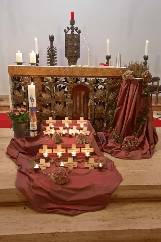 Vor einem Altar und der Osterkerze ist ein rotes Tuch ausgebreitet, darauf sind mehrere Holzkreuze ausgelegt mit Namen der Verstorbenen des letzten Jahres und Kerzen