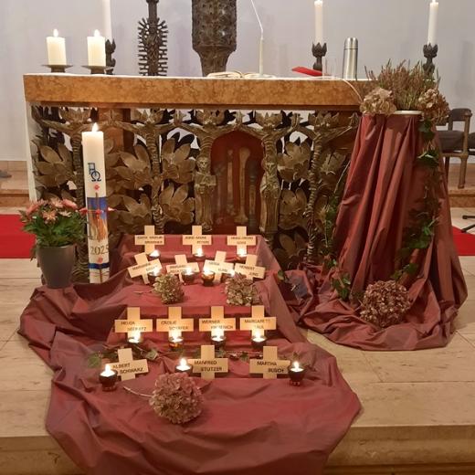 Vor einem Altar und der Osterkerze ist ein rotes Tuch ausgebreitet, darauf sind mehrere Holzkreuze ausgelegt mit Namen der Verstorbenen des letzten Jahres und Kerzen