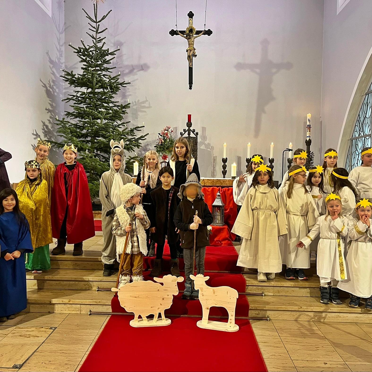 Kinder als Darsteller eines Krippenspiels gekleidet stehen im Altarraum einer Kirche, rechts eine Gruppe Engel, links und in der Mitte die Hirten, Tiere und die Heilige Familie. Im Hintergrund ein Weihnachtsbaum und ein Kreuz