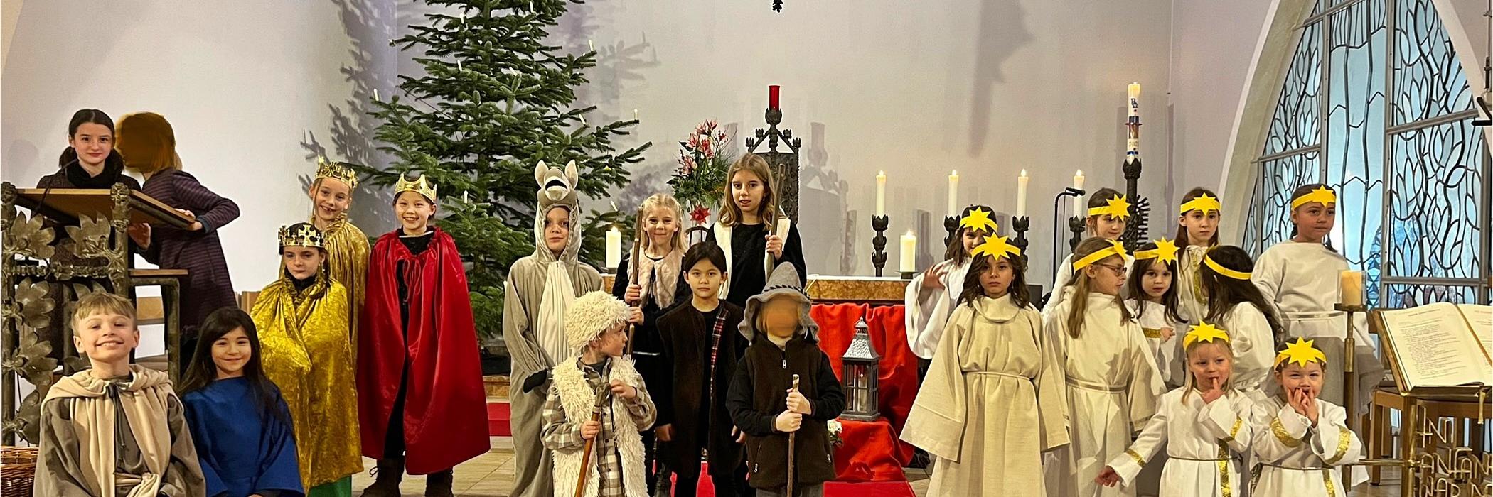 Kinder als Darsteller eines Krippenspiels gekleidet stehen im Altarraum einer Kirche, rechts eine Gruppe Engel, links und in der Mitte die Hirten, Tiere und die Heilige Familie. Im Hintergrund ein Weihnachtsbaum und ein Kreuz