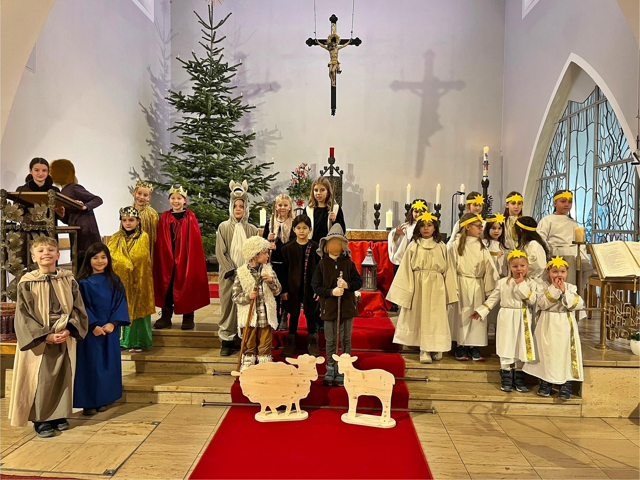 Kinder als Darsteller eines Krippenspiels gekleidet stehen im Altarraum einer Kirche, rechts eine Gruppe Engel, links und in der Mitte die Hirten, Tiere und die Heilige Familie. Im Hintergrund ein Weihnachtsbaum und ein Kreuz
