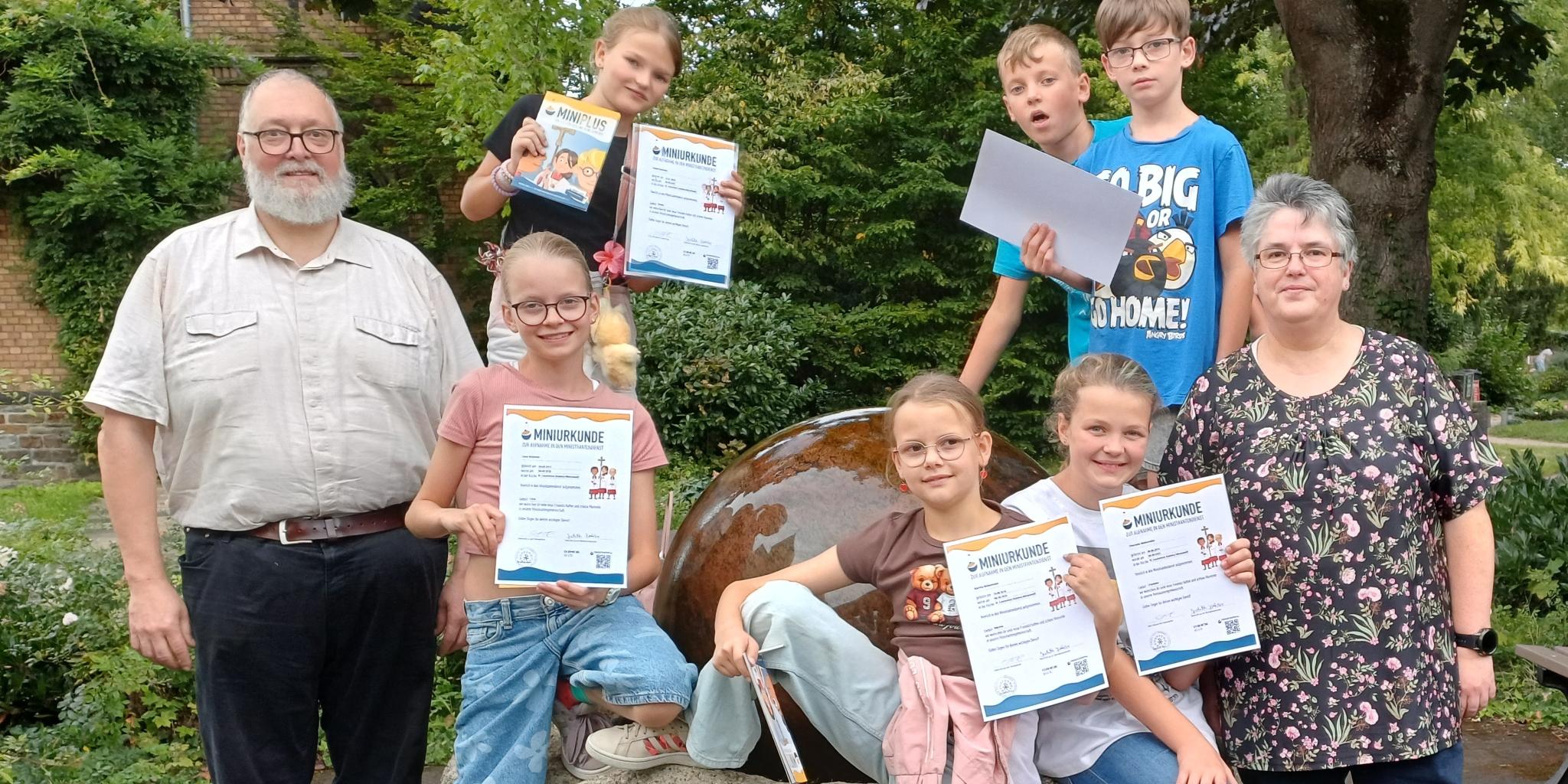 in der Mitte sechs Kinder, die eine Urkunde hochhalten, rechts und links stehen ein Mann und eine Frau
