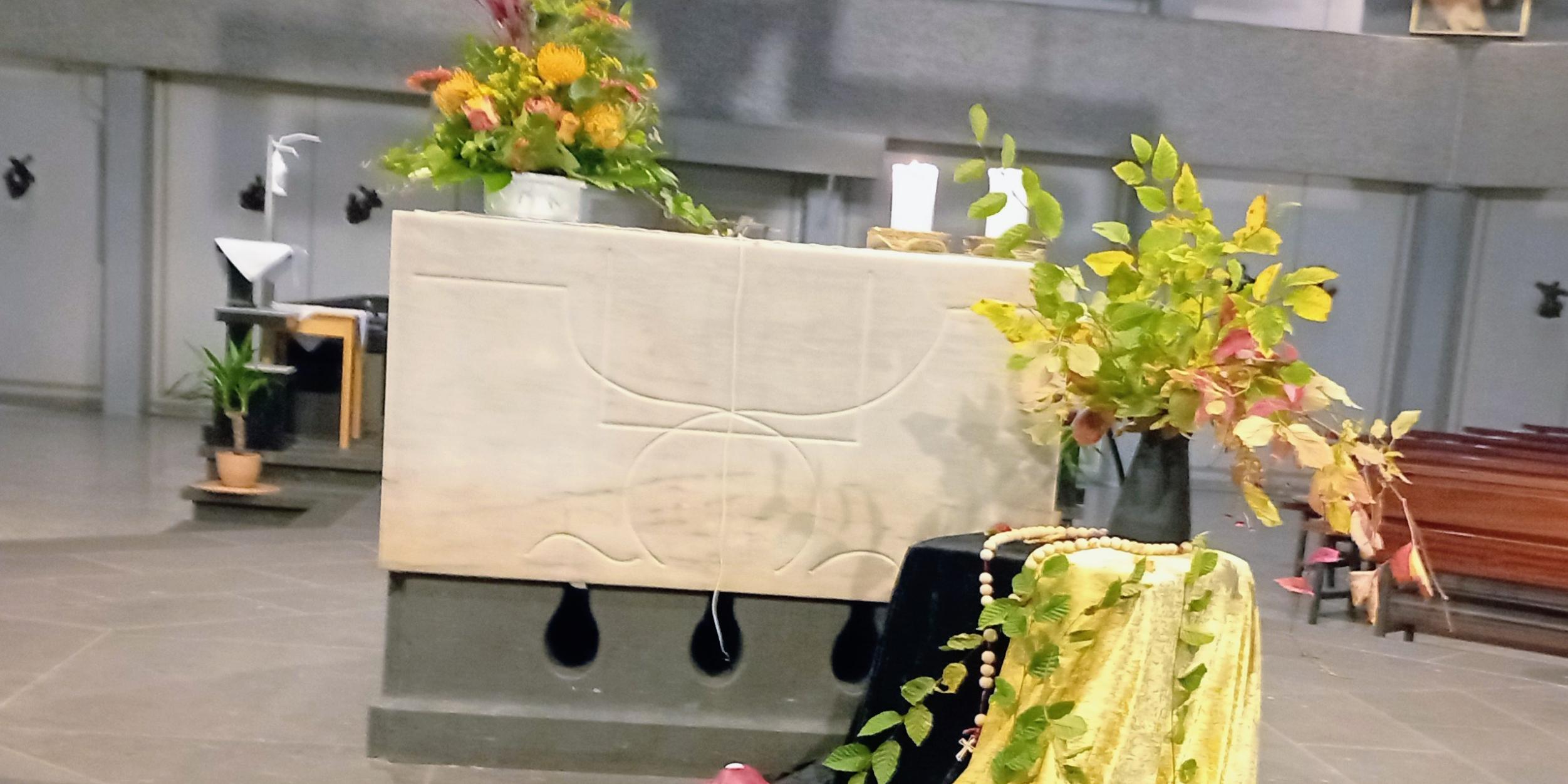 Rosenkranzandacht_St.Franziskus (c) Gaby Tampe Altar in der St. Franziskuskirche mit Sonnenblumen geschmückt davor ein Tisch, bedeckt mit einem schwarzen und gelben Tuch, darauf eine Vase mit herbstlichen Zweigen. Um die Vase ein großer Rosenkranz gelegt