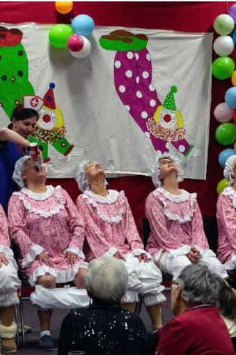 Foto eines Programmpunktes des Sitzung, auf der Bühne sitzen sechs Damen auf Stühlen in einer Reihe, diese sind bekleidet in altmodischen rosa-weißen Rüschenschlafanzügen, ein Dame erhält Sprühsahne in den Mund, ihre Hose ist in den Kniekehlen. Im Hintergrund die Bühnendeko bestehend aus Luftballons und einem Banner mit Clowns