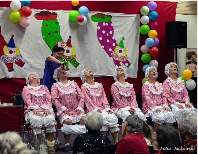 Foto eines Programmpunktes des Sitzung, auf der Bühne sitzen sechs Damen auf Stühlen in einer Reihe, diese sind bekleidet in altmodischen rosa-weißen Rüschenschlafanzügen, ein Dame erhält Sprühsahne in den Mund, ihre Hose ist in den Kniekehlen. Im Hintergrund die Bühnendeko bestehend aus Luftballons und einem Banner mit Clowns