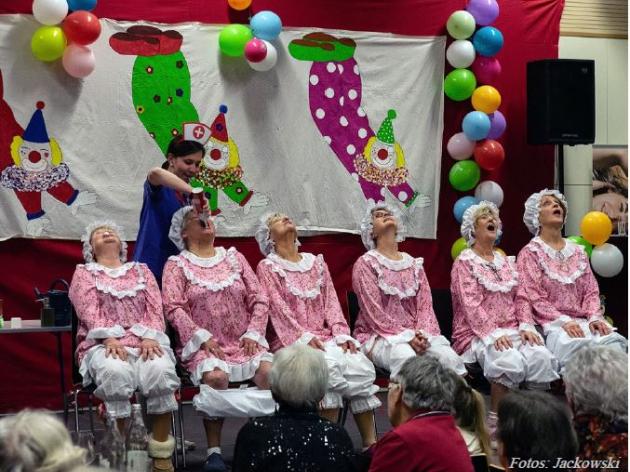 Foto eines Programmpunktes des Sitzung, auf der Bühne sitzen sechs Damen auf Stühlen in einer Reihe, diese sind bekleidet in altmodischen rosa-weißen Rüschenschlafanzügen, ein Dame erhält Sprühsahne in den Mund, ihre Hose ist in den Kniekehlen. Im Hintergrund die Bühnendeko bestehend aus Luftballons und einem Banner mit Clowns