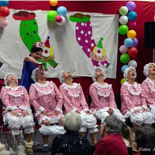 Foto eines Programmpunktes des Sitzung, auf der Bühne sitzen sechs Damen auf Stühlen in einer Reihe, diese sind bekleidet in altmodischen rosa-weißen Rüschenschlafanzügen, ein Dame erhält Sprühsahne in den Mund, ihre Hose ist in den Kniekehlen. Im Hintergrund die Bühnendeko bestehend aus Luftballons und einem Banner mit Clowns