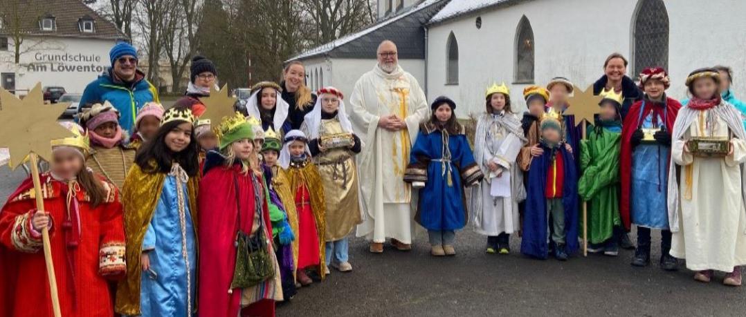 Eine Gruppe Kinder als Sternsinger gekleidet, in der Mitte Pfarrer Michael Frevel, im Hintergrund die Kirche St. Beatus
