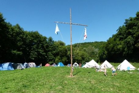 Zeltlager auf einer Wiese umrandet von Wald, in der Mitte ein Kreuz