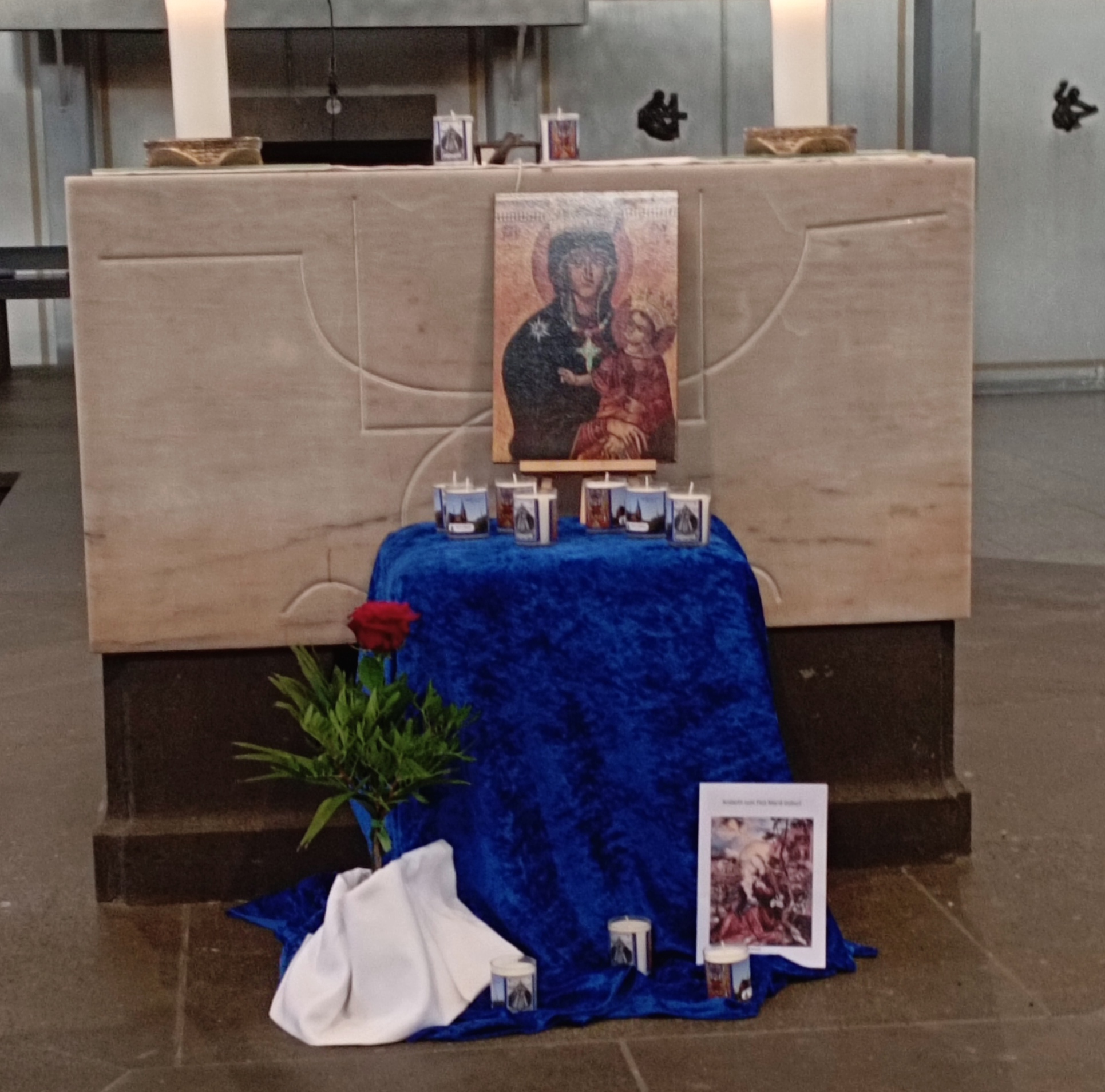 Vor dem Altar der St. Franziskuskirche steht ein Tisch, bedeckt mit blauem Tuch, darauf ein Bild der Muttergottes und Kerzen. Links vor dem Tisch eine Vase mit einer roten Rose, rechts daneben weitre Kerzen (c) Gaby Tampe Vor dem Altar der St. Franziskuskirche steht ein Tisch, bedeckt mit blauem Tuch, darauf ein Bild der Muttergottes und Kerzen. Links vor dem Tisch eine Vase mit einer roten Rose, rechts daneben weitre Kerzen