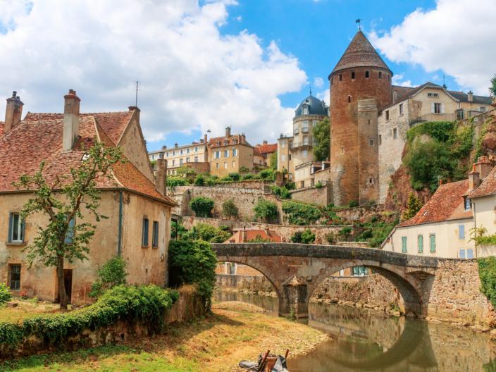 Zu sehen ist eine Ansicht der Stadt Semur, die  beidseitig an einem Fluss liegt über den eine mittelalterliche Brücke führt. Idylisch, malerisch sind mittelalterliche Gebäude und prominent ein Turm zu sehen.