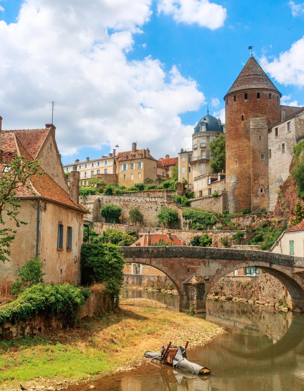 Zu sehen ist eine Ansicht der Stadt Semur, die  beidseitig an einem Fluss liegt über den eine mittelalterliche Brücke führt. Idylisch, malerisch sind mittelalterliche Gebäude und prominent ein Turm zu sehen.