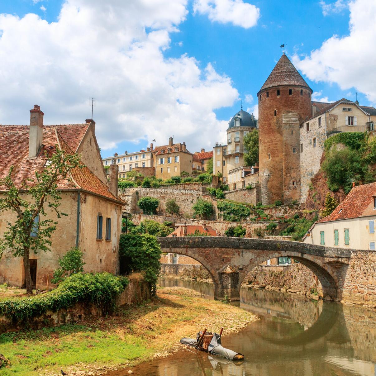 Zu sehen ist eine Ansicht der Stadt Semur, die  beidseitig an einem Fluss liegt über den eine mittelalterliche Brücke führt. Idylisch, malerisch sind mittelalterliche Gebäude und prominent ein Turm zu sehen.