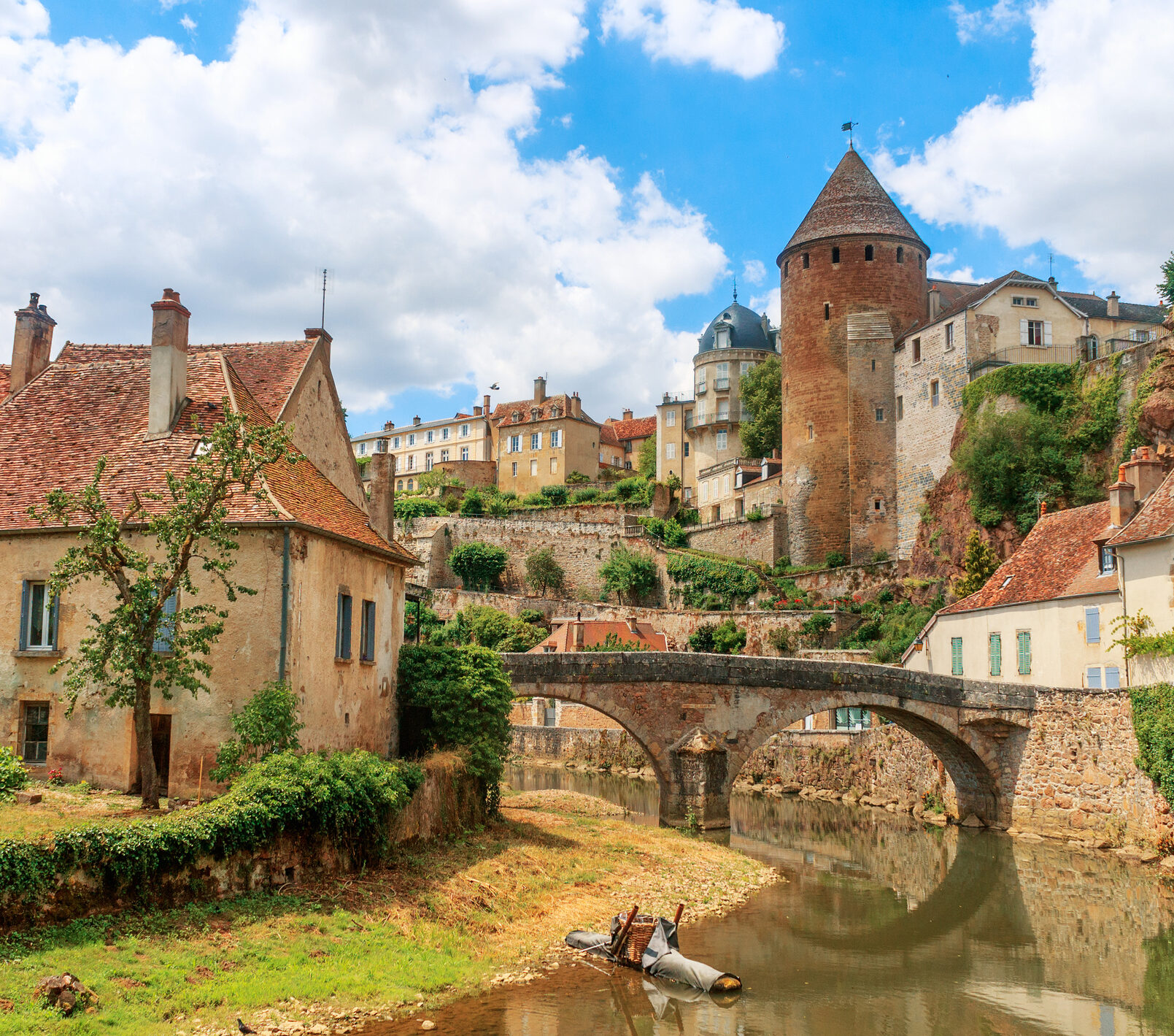 Zu sehen ist eine Ansicht der Stadt Semur, die  beidseitig an einem Fluss liegt über den eine mittelalterliche Brücke führt. Idylisch, malerisch sind mittelalterliche Gebäude und prominent ein Turm zu sehen.