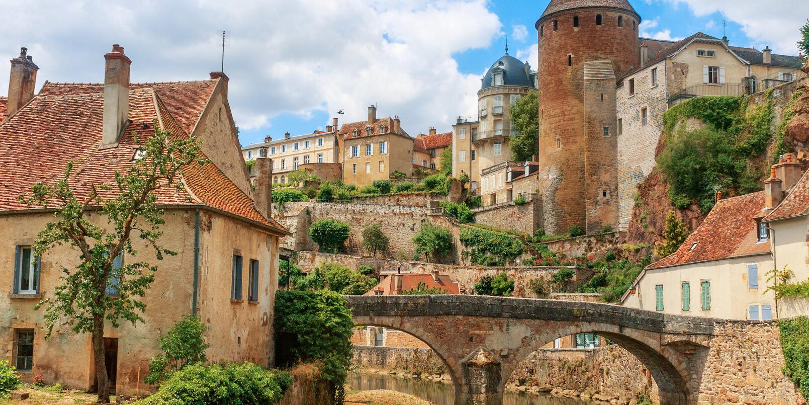 Zu sehen ist eine Ansicht der Stadt Semur, die  beidseitig an einem Fluss liegt über den eine mittelalterliche Brücke führt. Idylisch, malerisch sind mittelalterliche Gebäude und prominent ein Turm zu sehen.