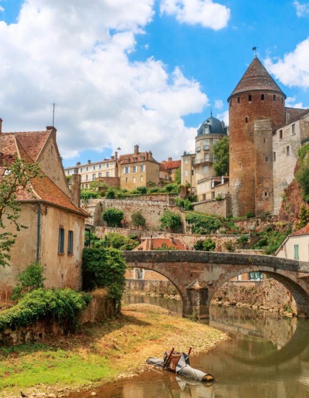 Zu sehen ist eine Ansicht der Stadt Semur, die  beidseitig an einem Fluss liegt über den eine mittelalterliche Brücke führt. Idylisch, malerisch sind mittelalterliche Gebäude und prominent ein Turm zu sehen.