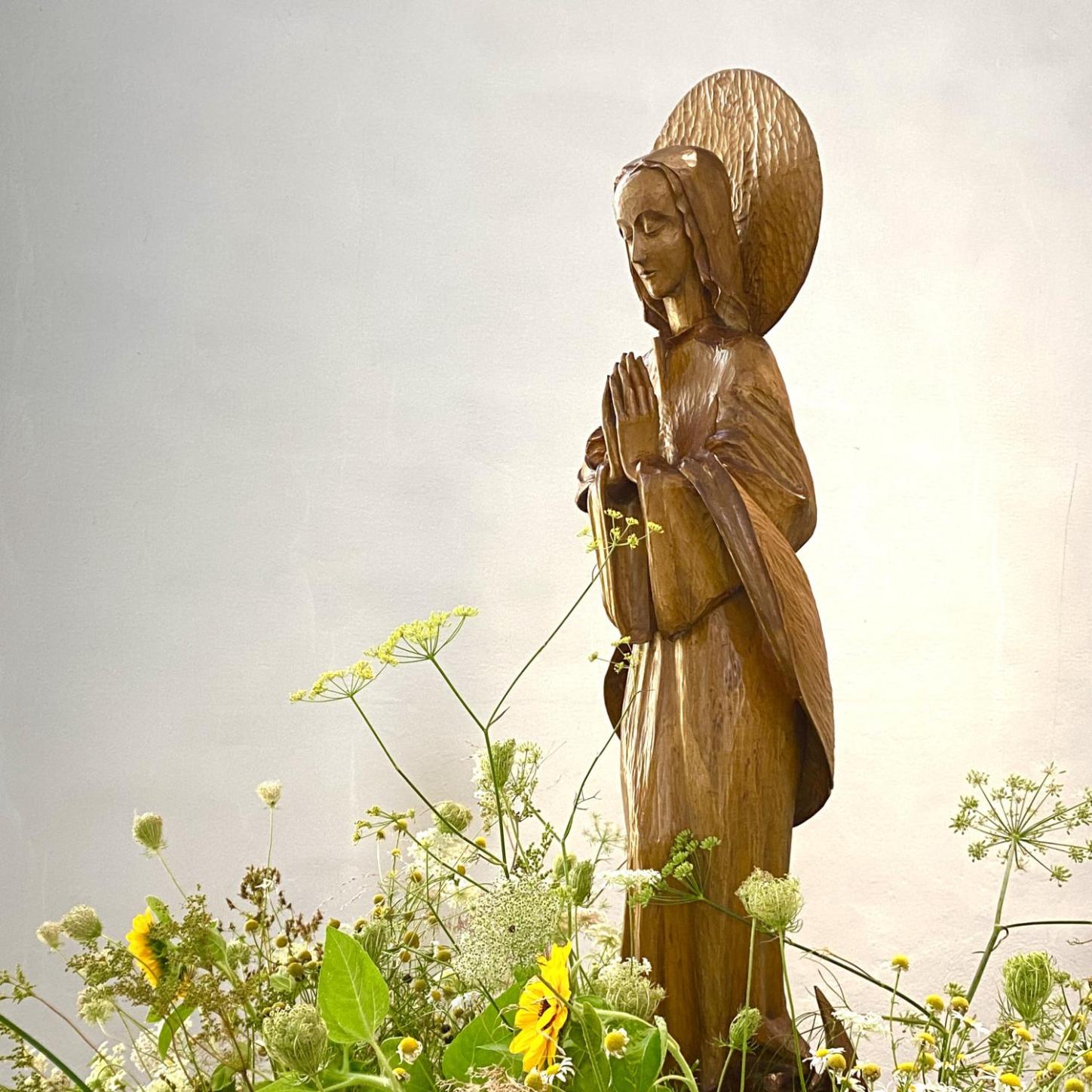 Holzstatue der betenden Gottesmutter Maria, umgeben von bunten Blumen und Gräsern.
