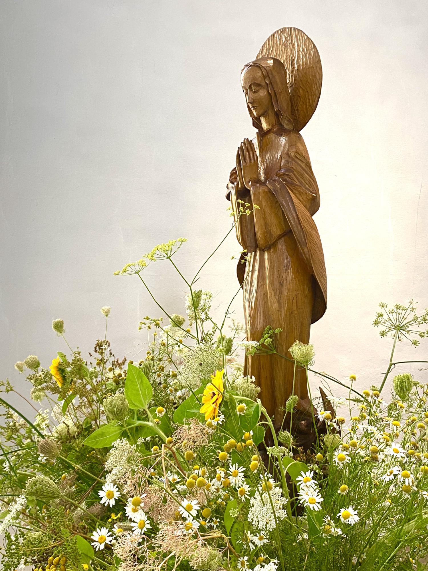 Holzstatue der betenden Gottesmutter Maria, umgeben von bunten Blumen und Gräsern.