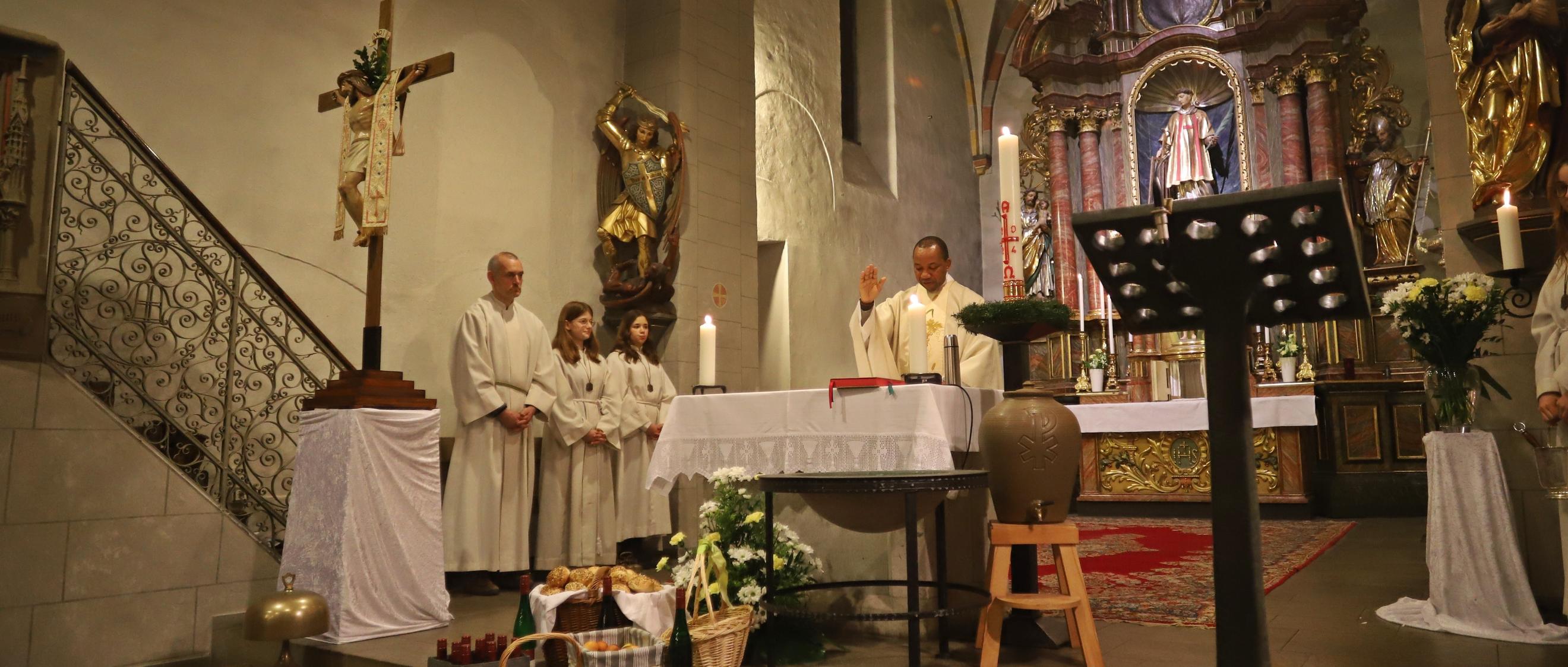 Blick in den Altarraum in der Osternacht, im Vordergrund das Kreuz und die Ostergaben, im Hintergrund drei Meßdiener und Pater segnet