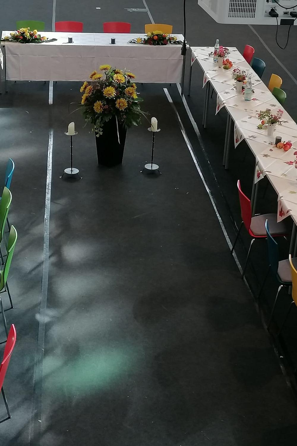 gedeckte und blumengeschmückte Tische als u-förmige Tafel aufgestellt, mit bunten Stühlen, in der Mitte eine große Blumenstaude und rechts und links je ein Kerzenständer